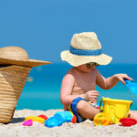 海辺の砂浜で砂遊びをする赤ちゃんの画像。帽子とサングラスをつけ、赤ちゃんでも使える日焼け止め（サンスクリーン剤）を塗って紫外線対策をおこなっている。