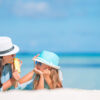 海で日差しの中、帽子をかぶり、紫外線対策をしている親子の画像。紫外線対策として、子供、赤ちゃんにおすすめの日焼け止めスティック、日焼け止めクリームを持っている。