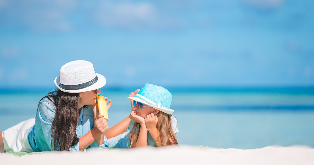 海で日差しの中、帽子をかぶり、紫外線対策をしている親子の画像。紫外線対策として、子供、赤ちゃんにおすすめの日焼け止めスティック、日焼け止めクリームを持っている。
